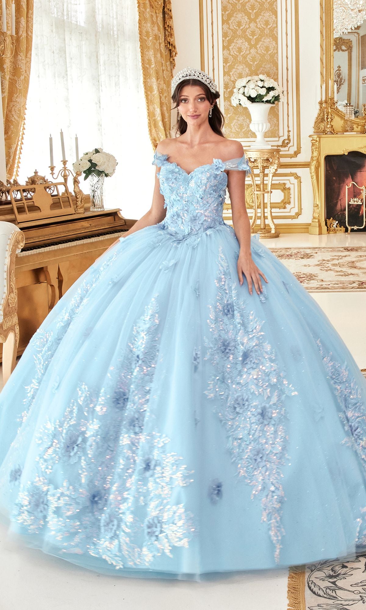 A woman wearing the Quinceanera Dress 15713 by Ladivine, a light blue off-the-shoulder gown with butterfly appliques and a tiara, stands in an ornate, gold-accented room.
